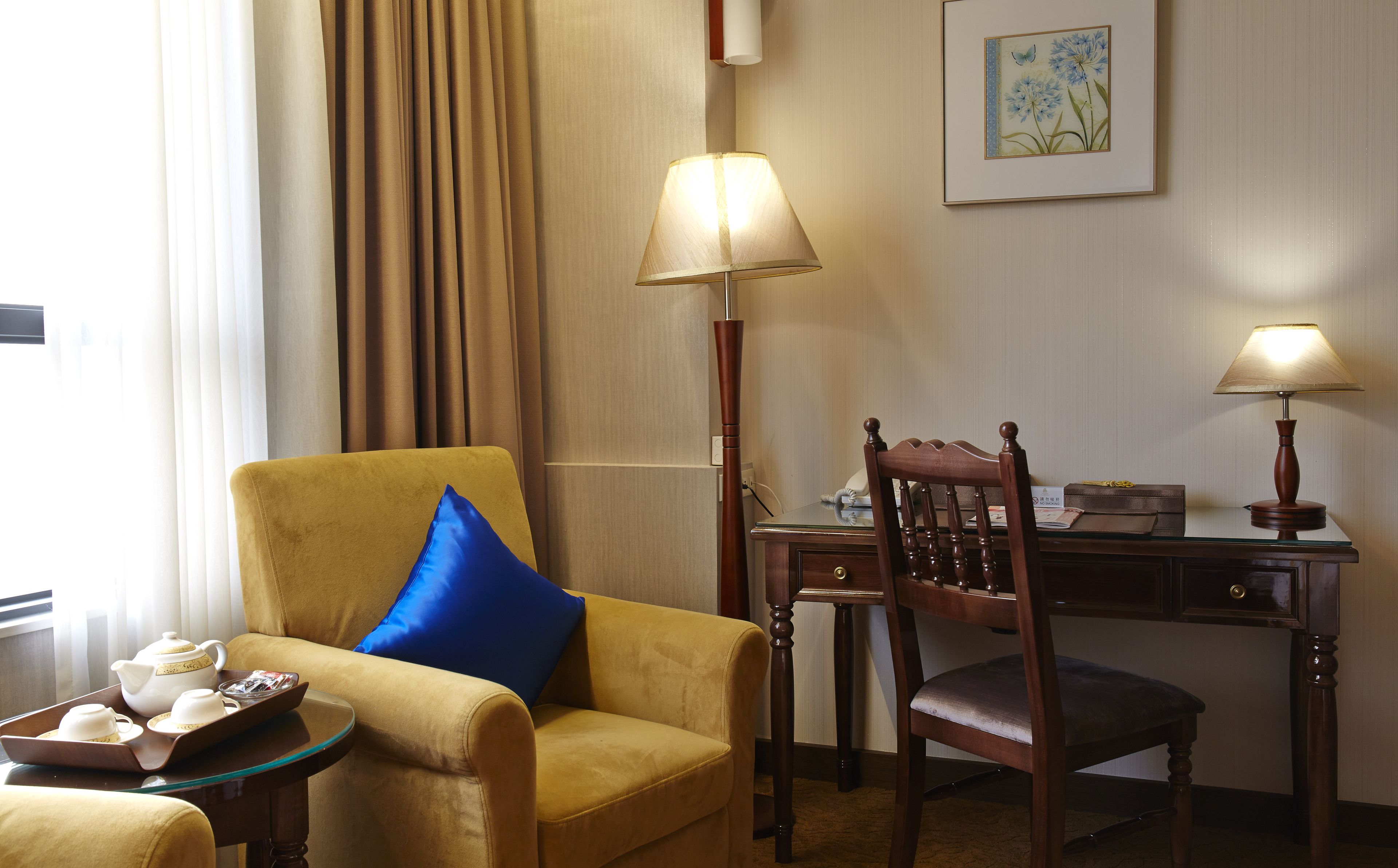 A hotel room with a yellow armchair, a wooden chair, a desk with a lamp, and a tray with teacups and saucers.