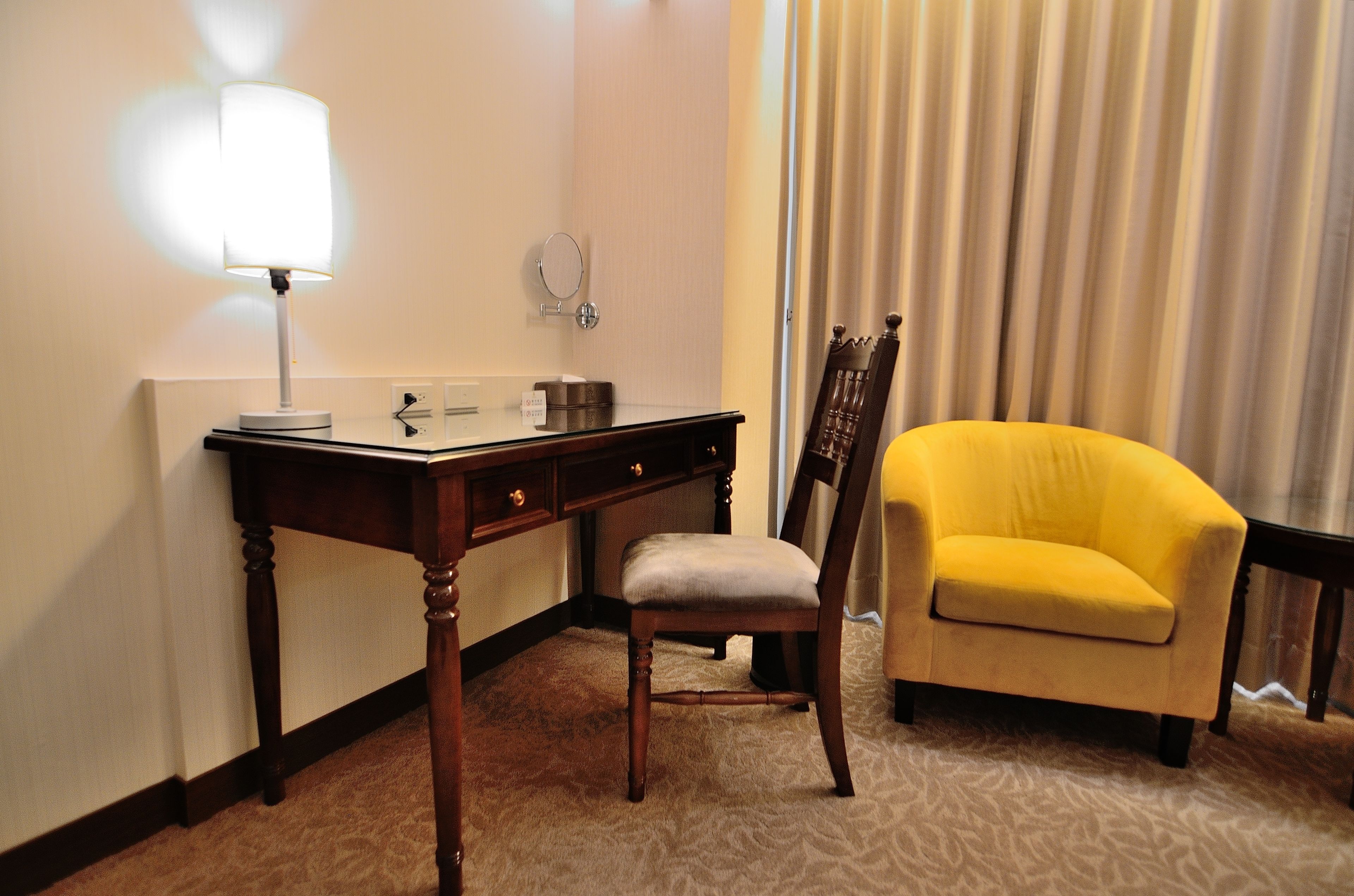 A hotel room with a desk, a chair, a lamp, a mirror, and a window with curtains.