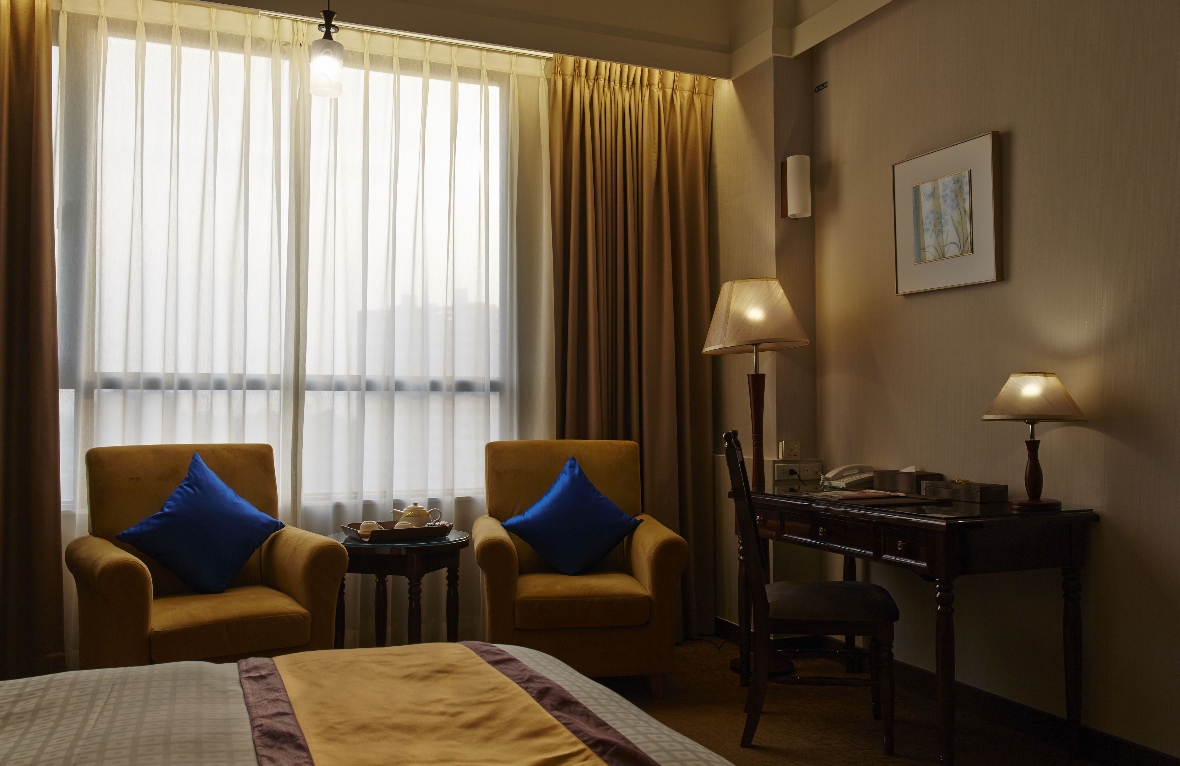 A hotel room with a bed, two armchairs with blue cushions, a desk with a lamp, and a window with sheer curtains.