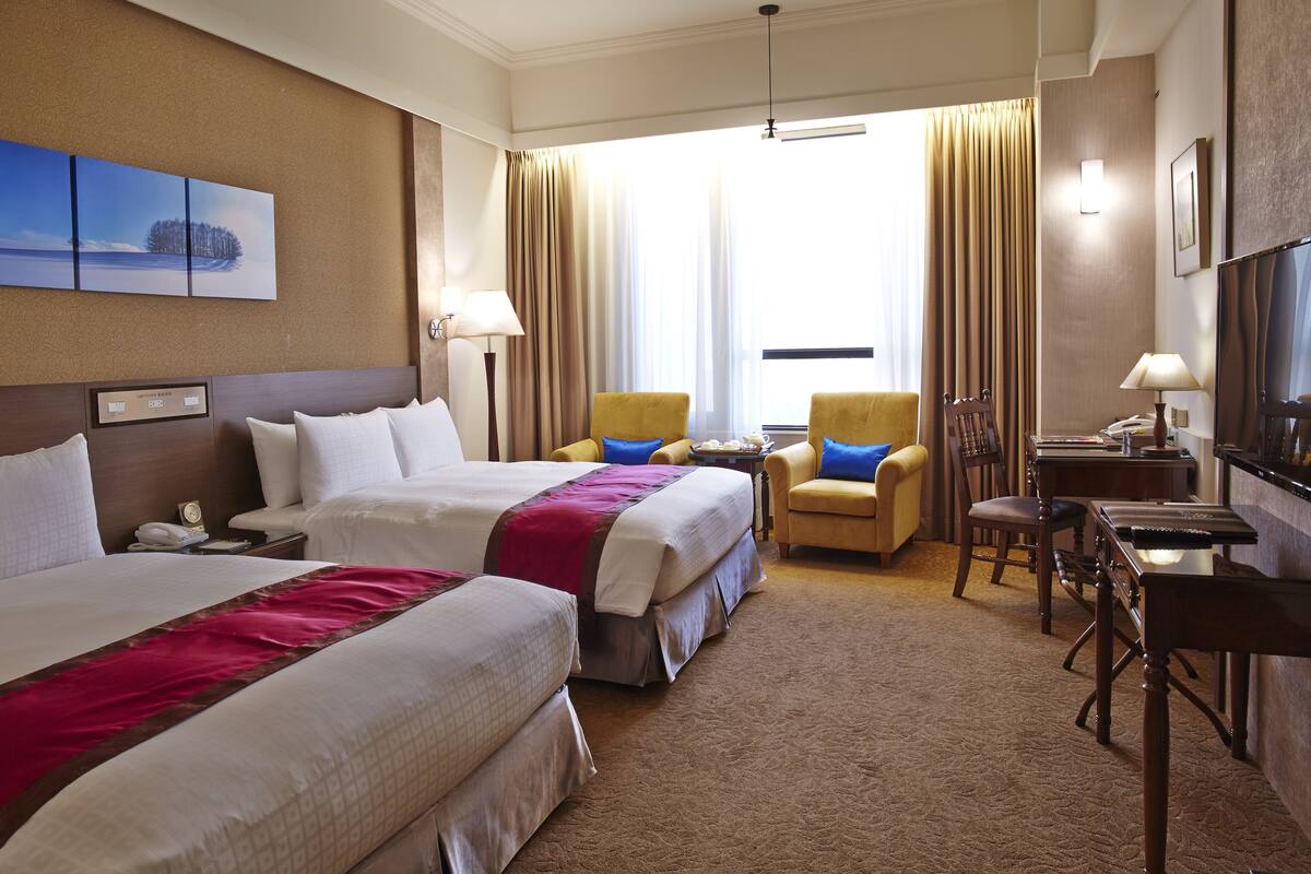 A hotel room with two beds, a desk, a chair, a TV, and a window with curtains.