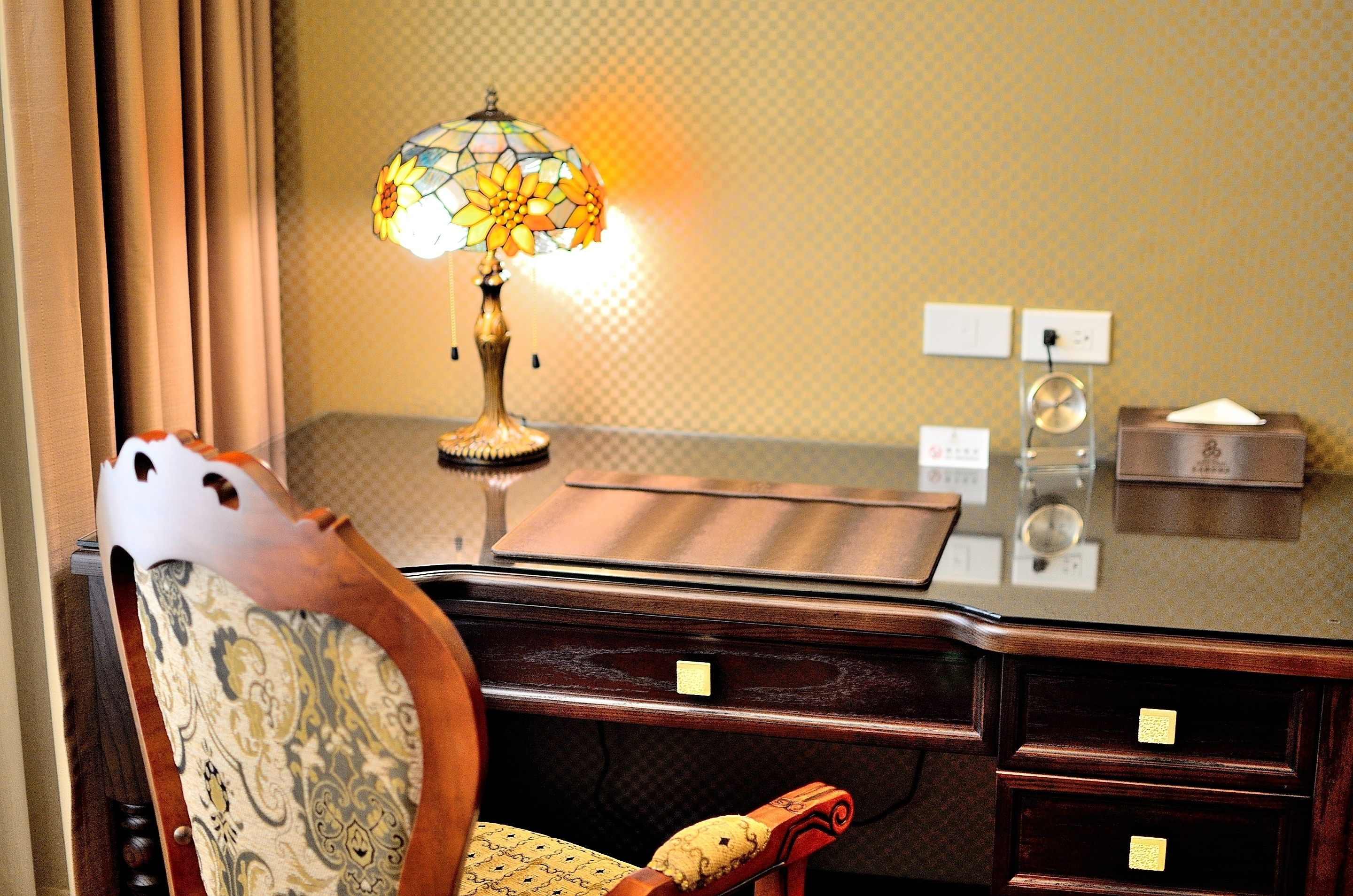 A wooden desk with a stained glass lamp, a chair with a patterned cushion, and a small wooden box on the desk.