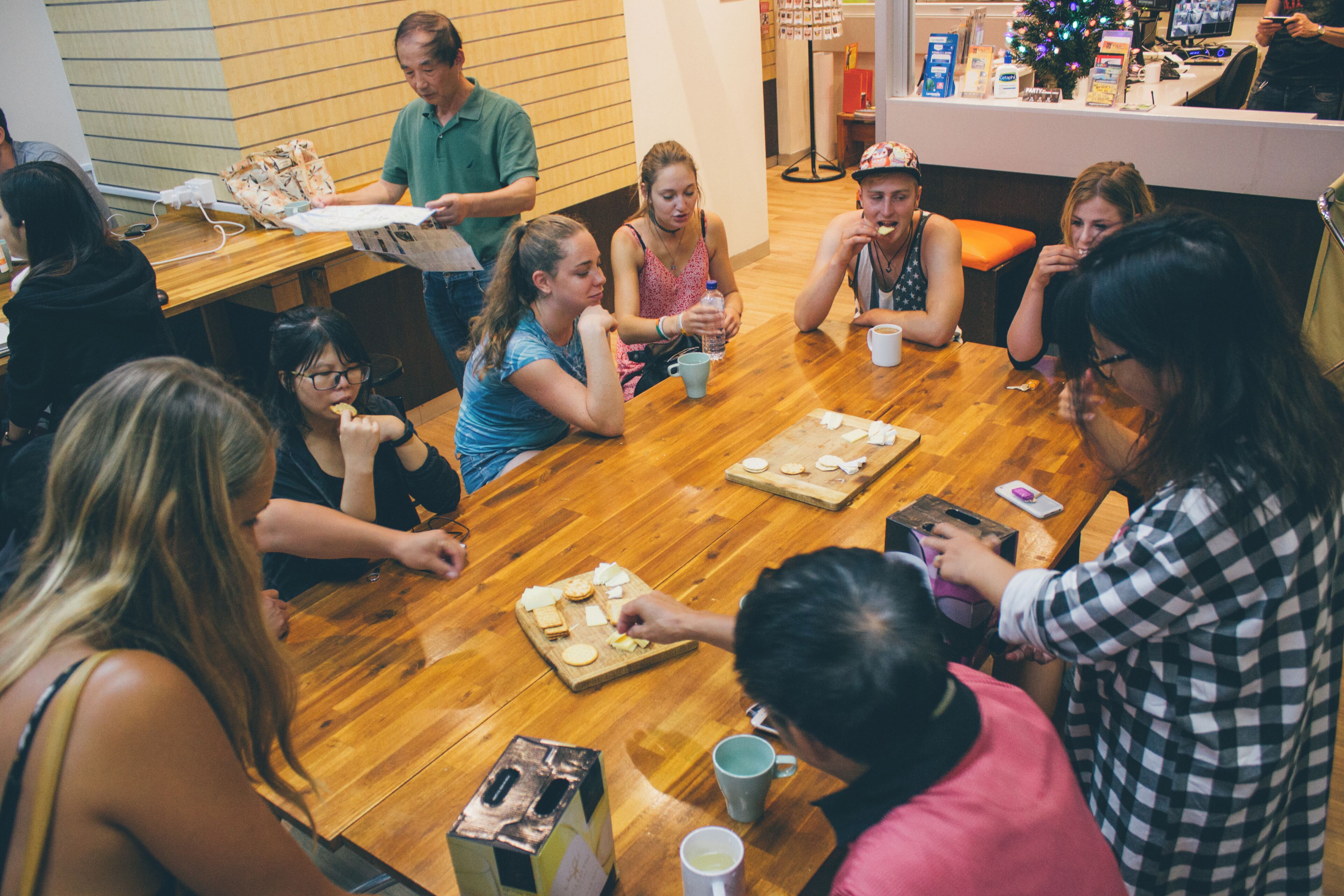A group of people sitting around a wooden table in a room with a tiled wall and a counter with various items.