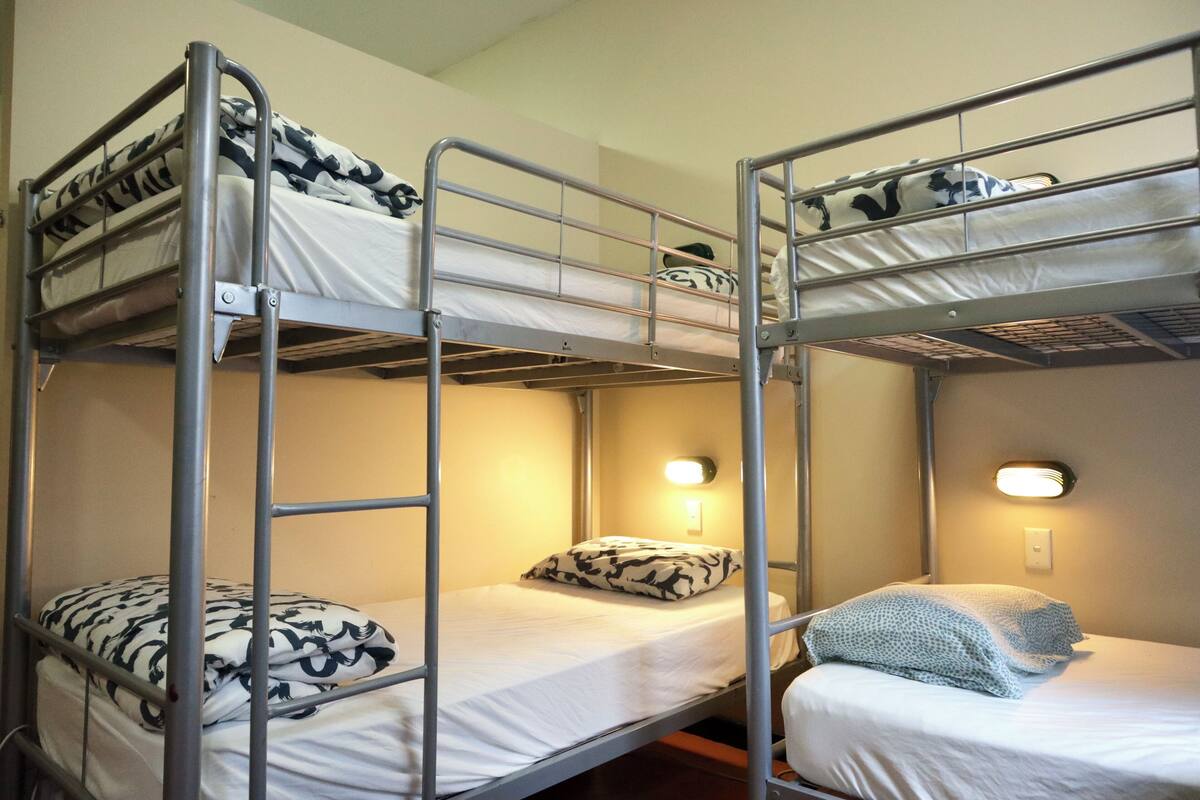 A bunk bed room with a metal frame, two beds with patterned bedding, a wall-mounted light, and a power outlet.