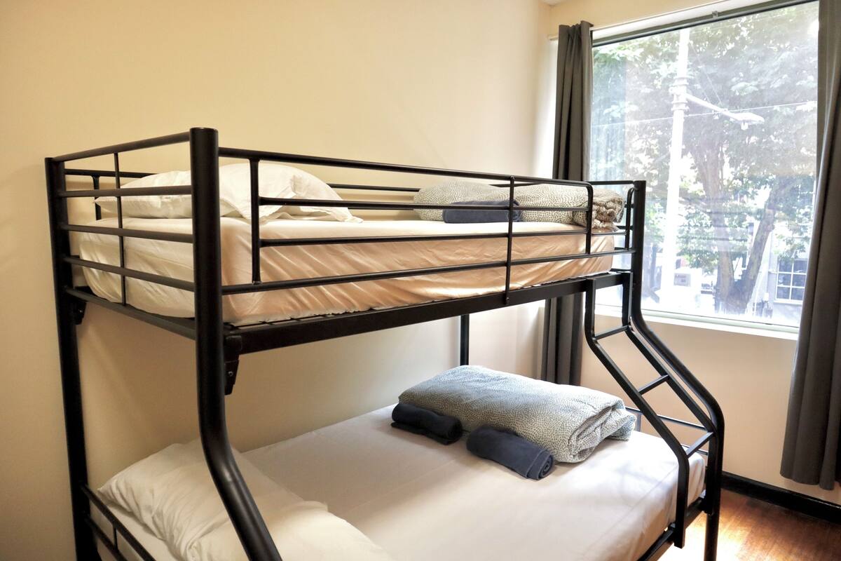 A bunk bed with a lower bunk, a window with curtains, and a view of trees outside.