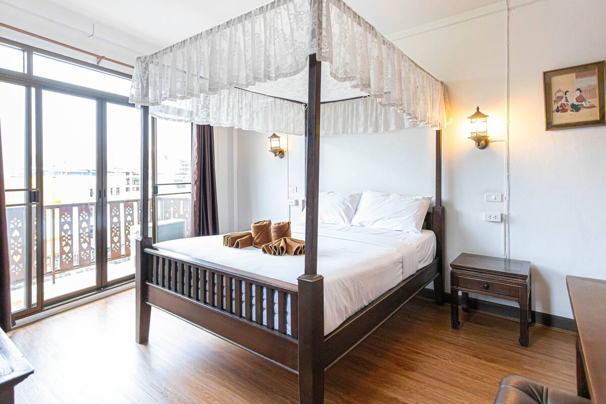 Una cama con dosel sobre cuatro columnas, con sábanas blancas, suelo de madera, una mesita de noche pequeña y una lámpara fijada a la pared.