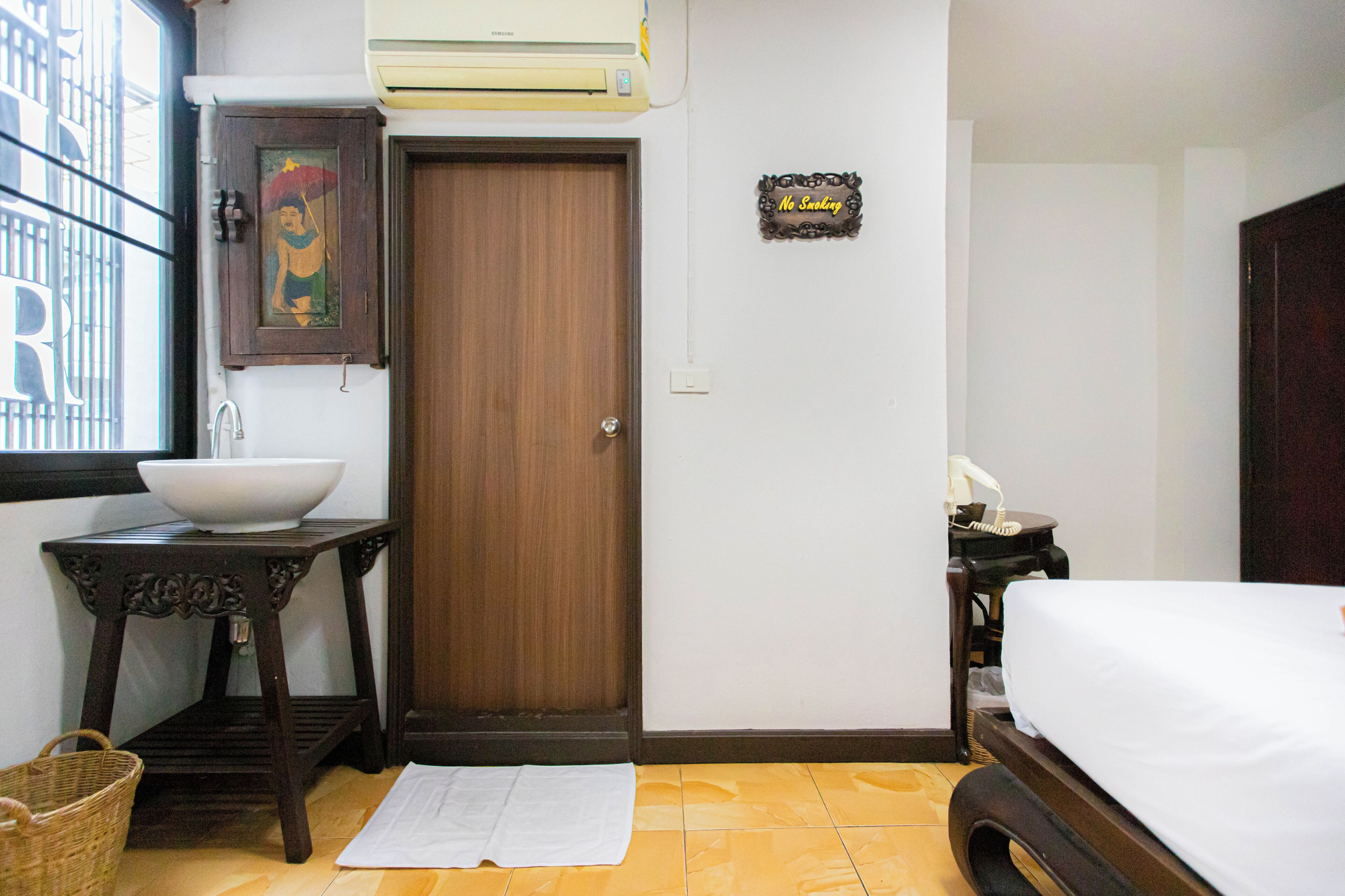 A hotel room with a wooden door, a white sink, a small table with a bowl, a bed, and a 'No Smoking' sign.
