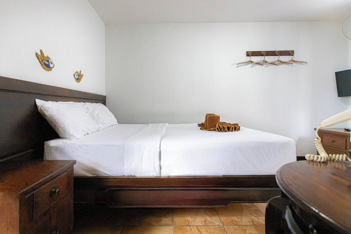 Una cama bien tendida con sábanas blancas, una mesita de noche de madera con cajón, un aparador de madera y un perchero con colgadores en la pared.