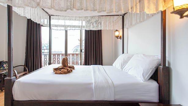Een opgemaakt bed met witte lakens en een bruine handdoek, een houten hoofdbord en een raam met gordijnen.