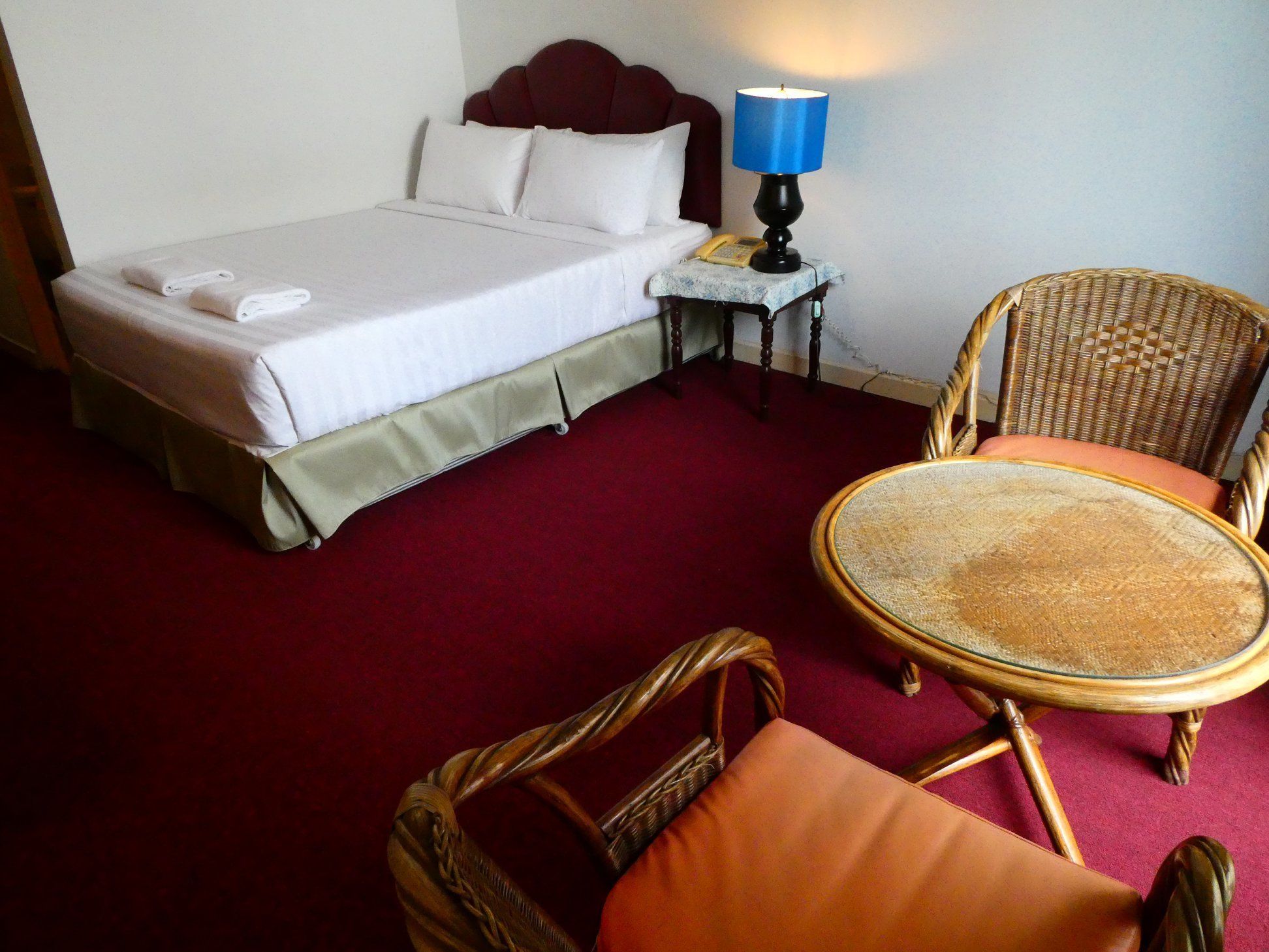 A hotel room with a bed, a small table with a lamp, and two wicker chairs.
