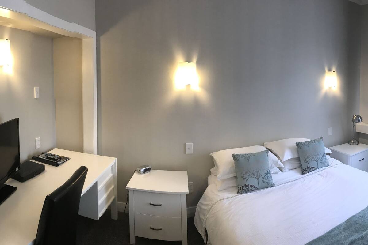 Una habitación de hotel con cama, escritorio con televisor, silla, mesita de noche y lámpara.