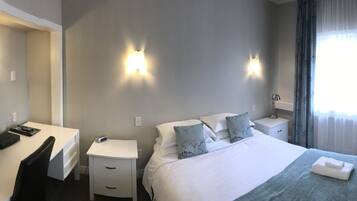 A hotel room with a bed, a desk with a TV, a chair, a nightstand, and a lamp.
