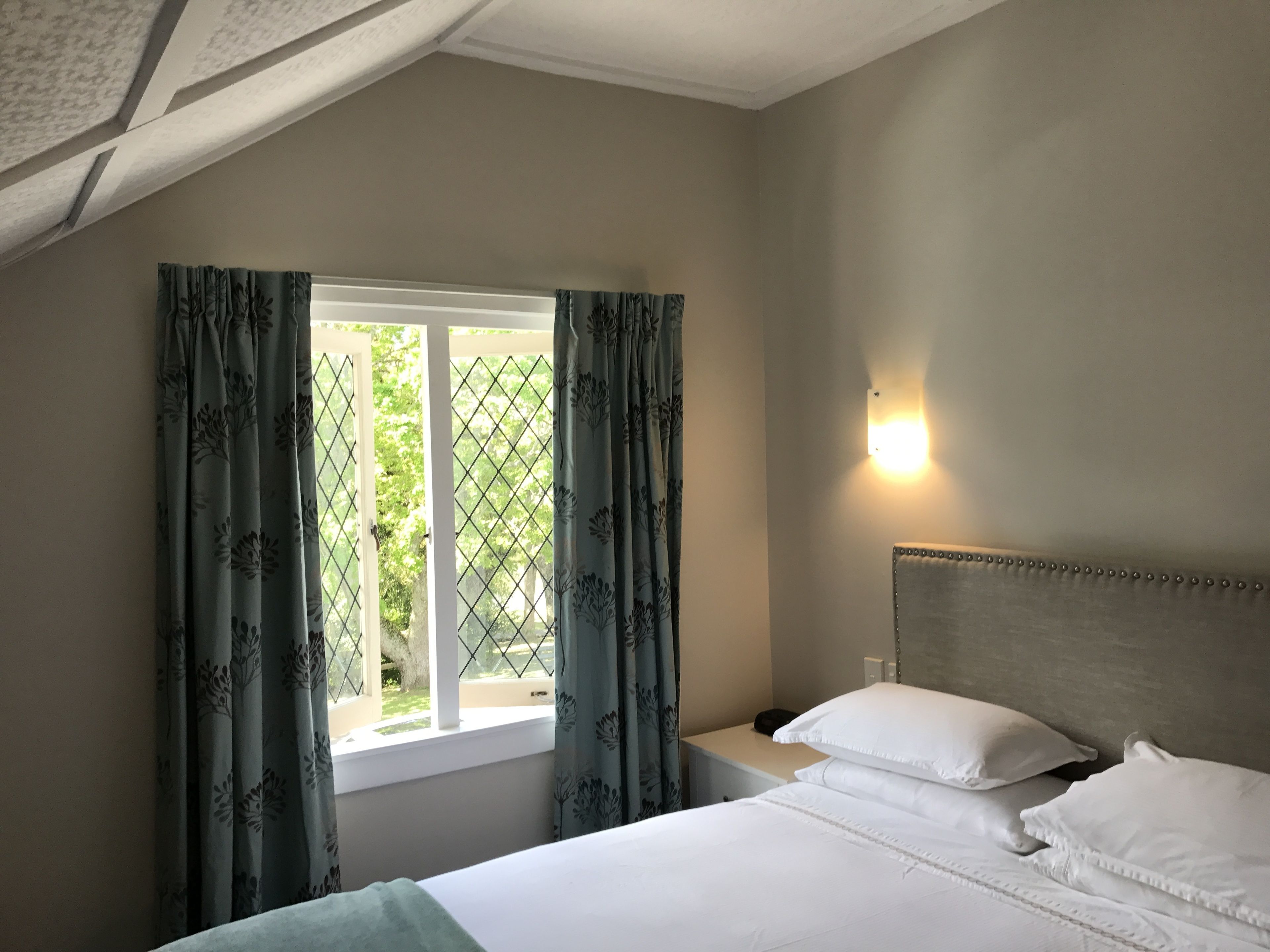 A bedroom with a bed, white pillows, a nightstand, a window with curtains, and a wall-mounted light.