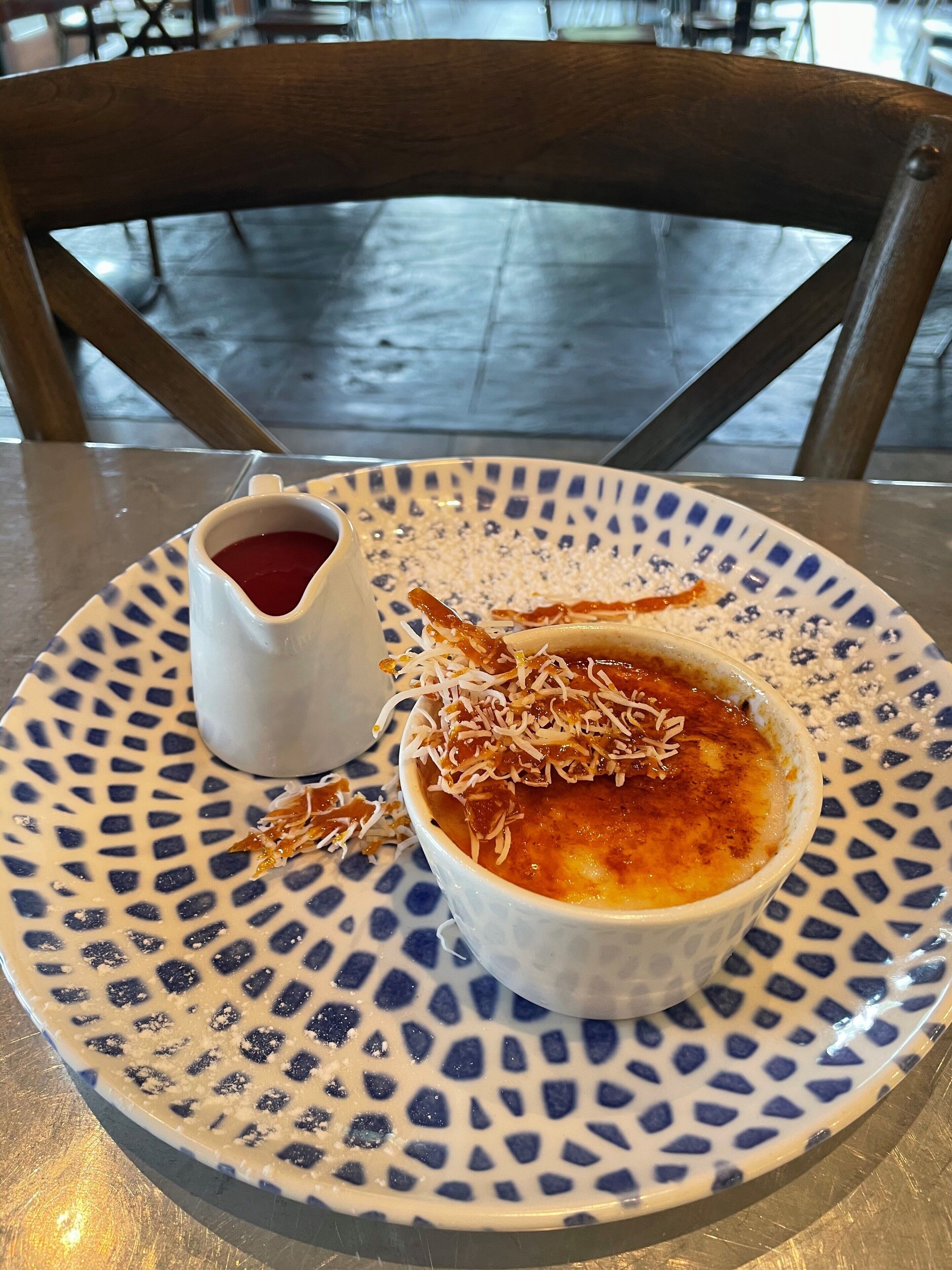 A dessert plate with a crème brûlée and a small cup of sauce, placed on a table with a patterned tablecloth.