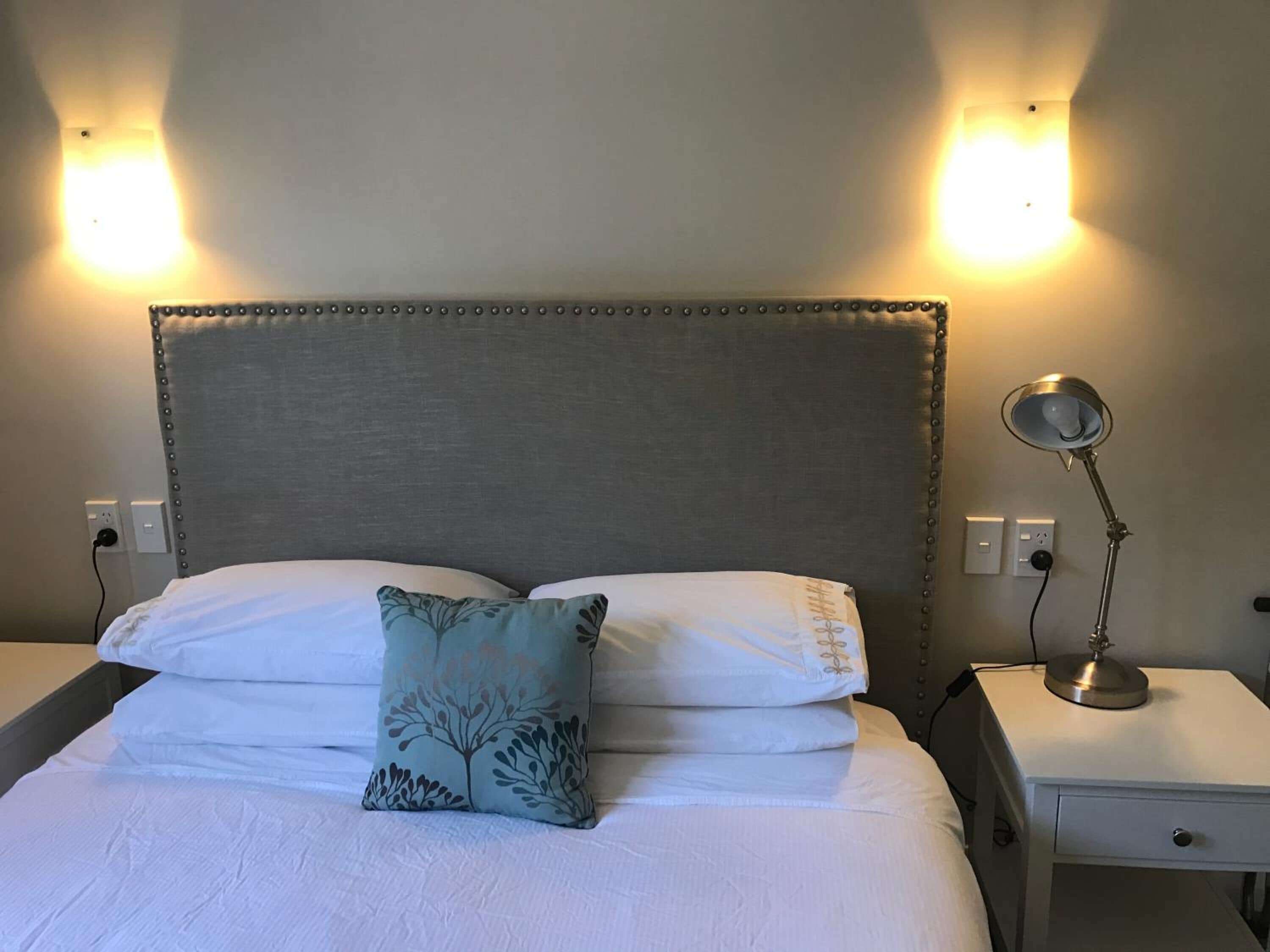 A neatly made bed with white linens and a decorative pillow, flanked by two bedside tables with lamps, against a plain wall with two wall-mounted lights.