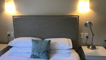 A neatly made bed with white linens and a decorative pillow, flanked by two bedside tables with lamps, against a plain wall with two wall-mounted lights.