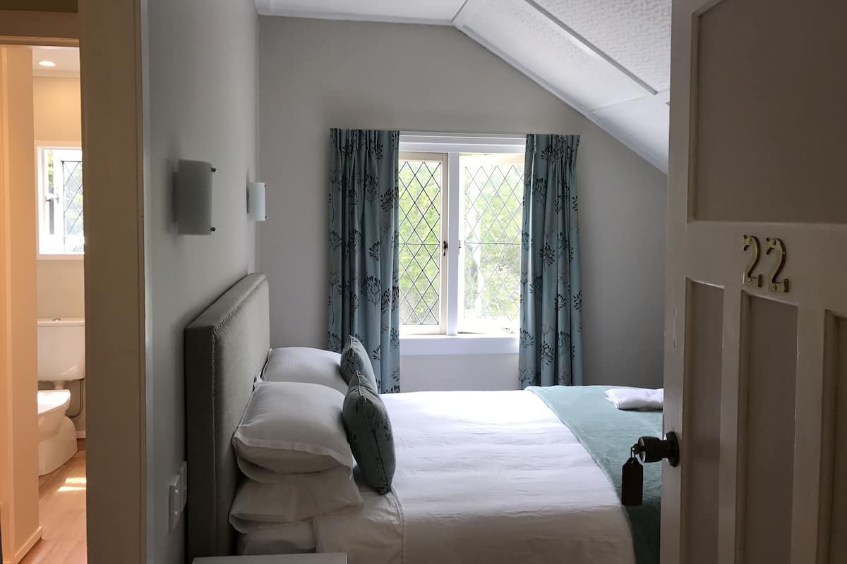 Un dormitorio con cama, ventana con cortinas, puerta con un número y vista al baño.