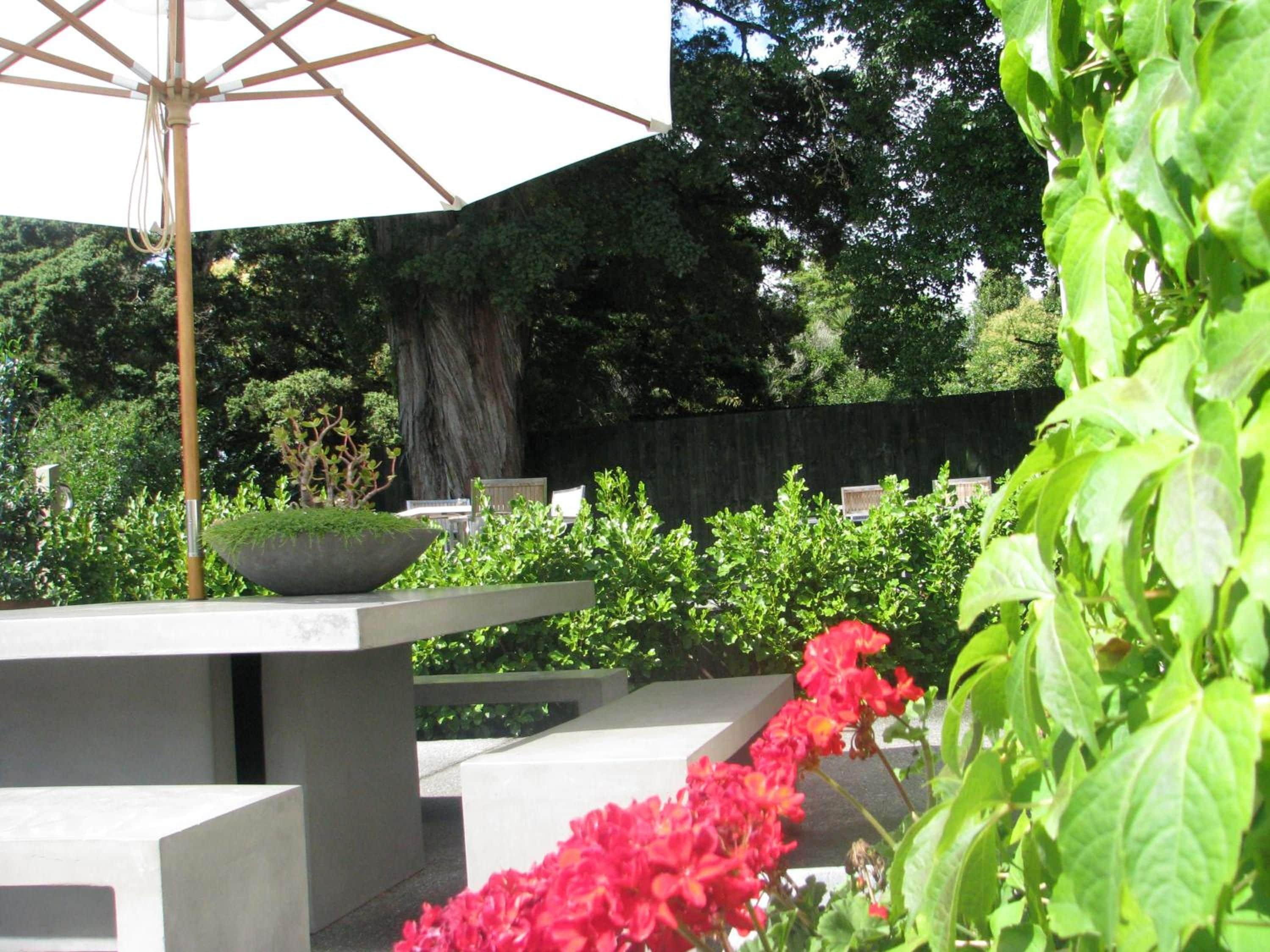 An outdoor seating area with a concrete table and chairs, surrounded by greenery and a large umbrella.