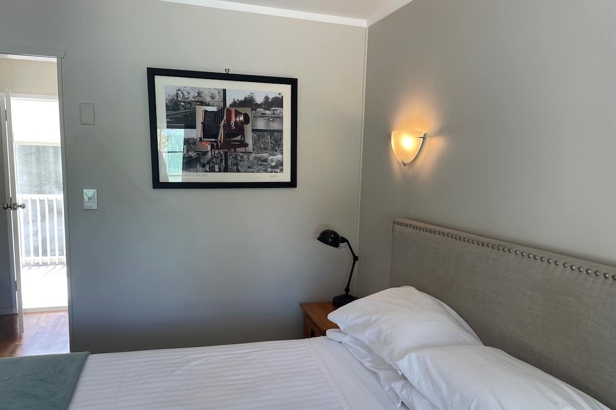 Un dormitorio con una cama, una fotografía enmarcada en la pared, una mesita de noche con lámpara y una puerta que da a un balcón.