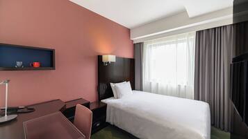 A hotel room with a bed, desk, chair, and a small shelf with a cup and mug.