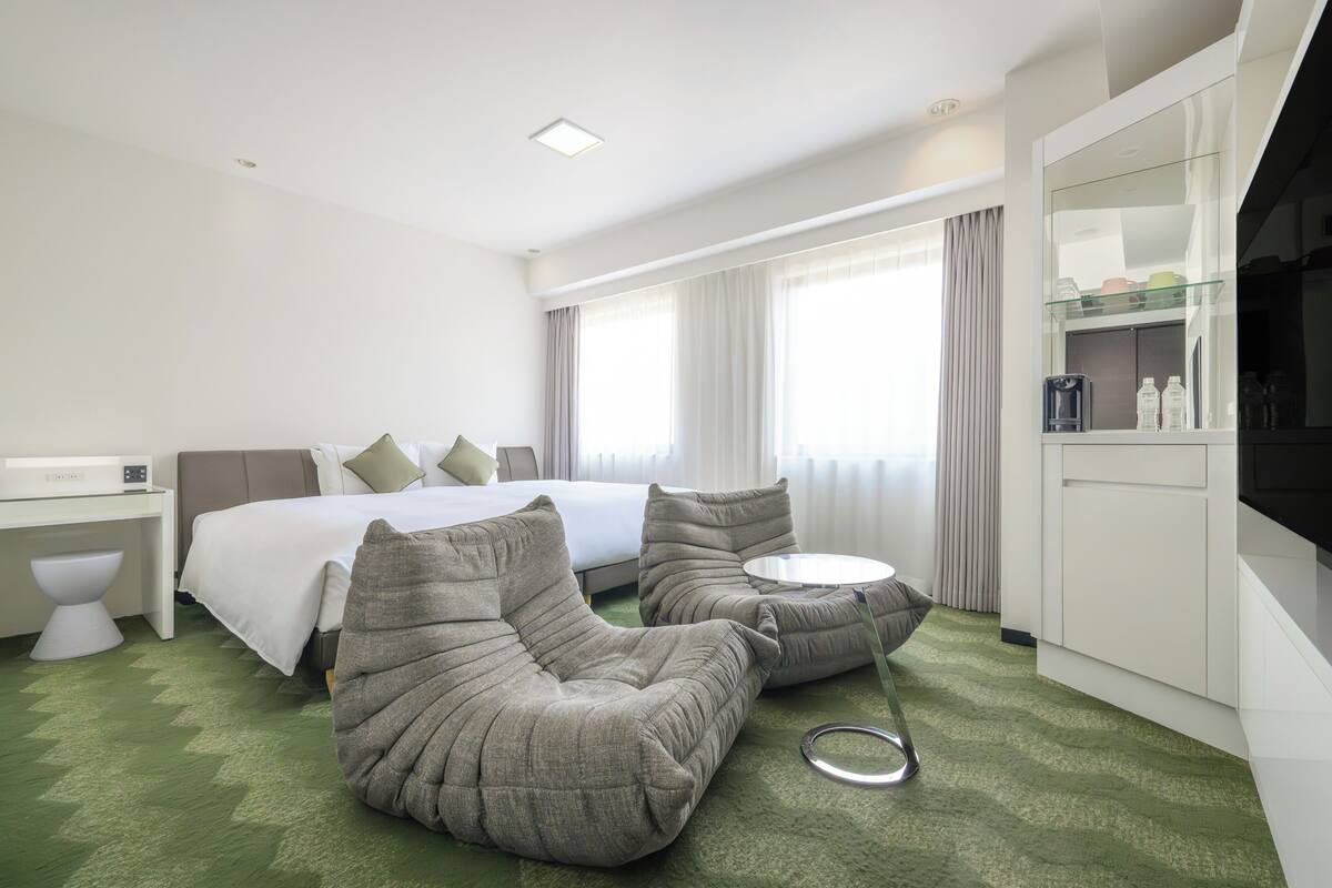 A hotel room with a bed, two grey sofas, a small round table, a white desk, a white toilet, and a large mirror.