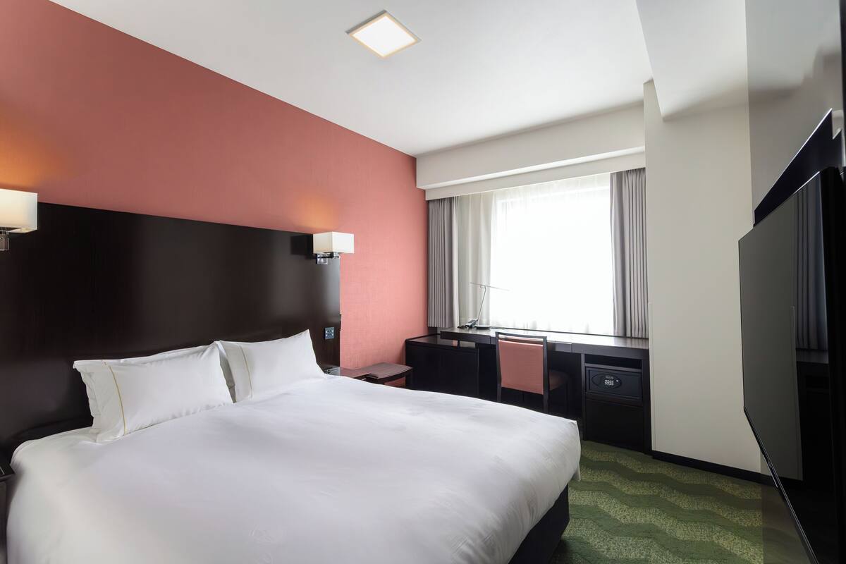 A hotel room with a large bed, a desk with a chair, a TV, and a window with curtains.