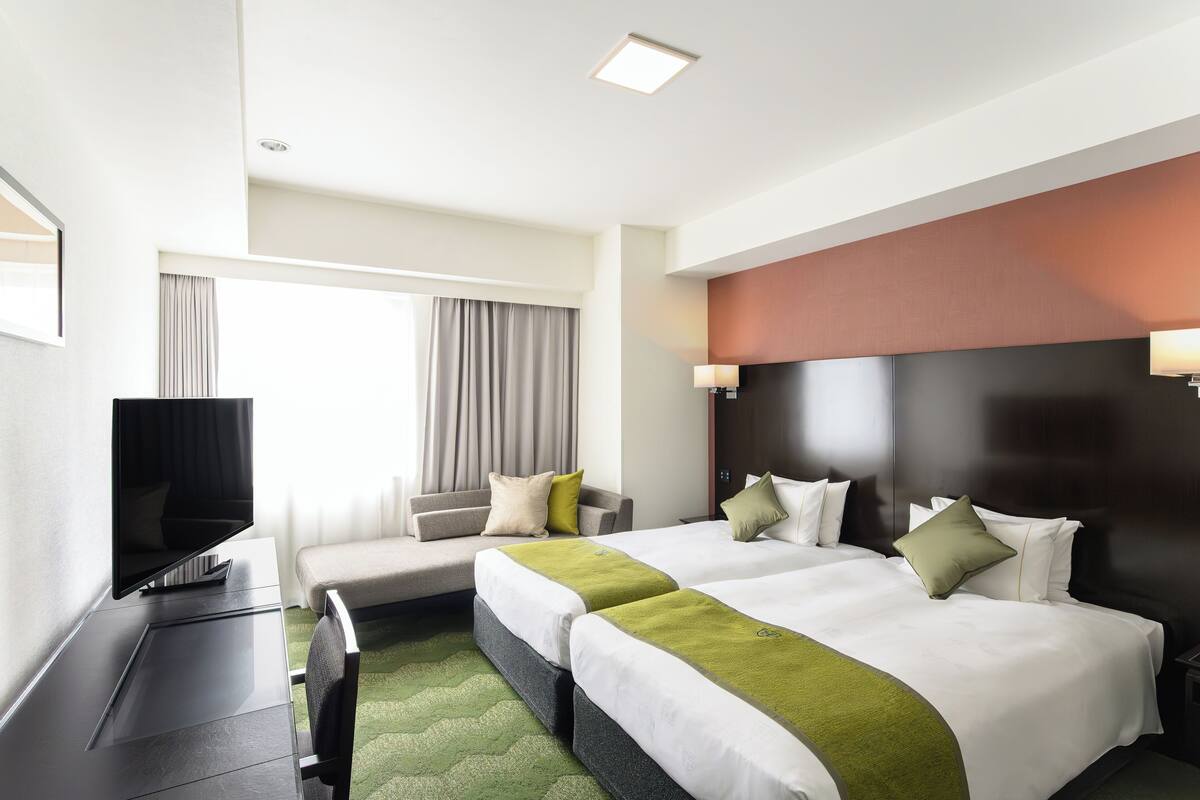A hotel room with two beds, a desk, a TV, and a window with curtains.