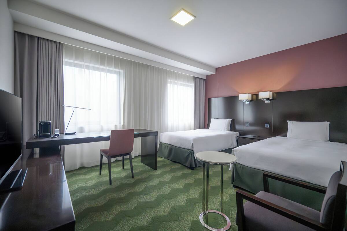 A hotel room with two beds, a desk, a chair, a small table, and a TV.