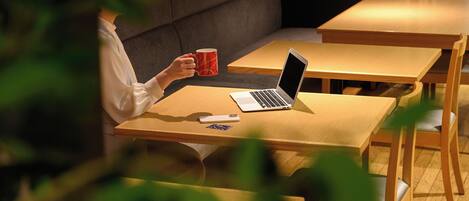 A person sitting at a wooden table with a laptop and a red mug, in a room with a sofa and another table in the background.