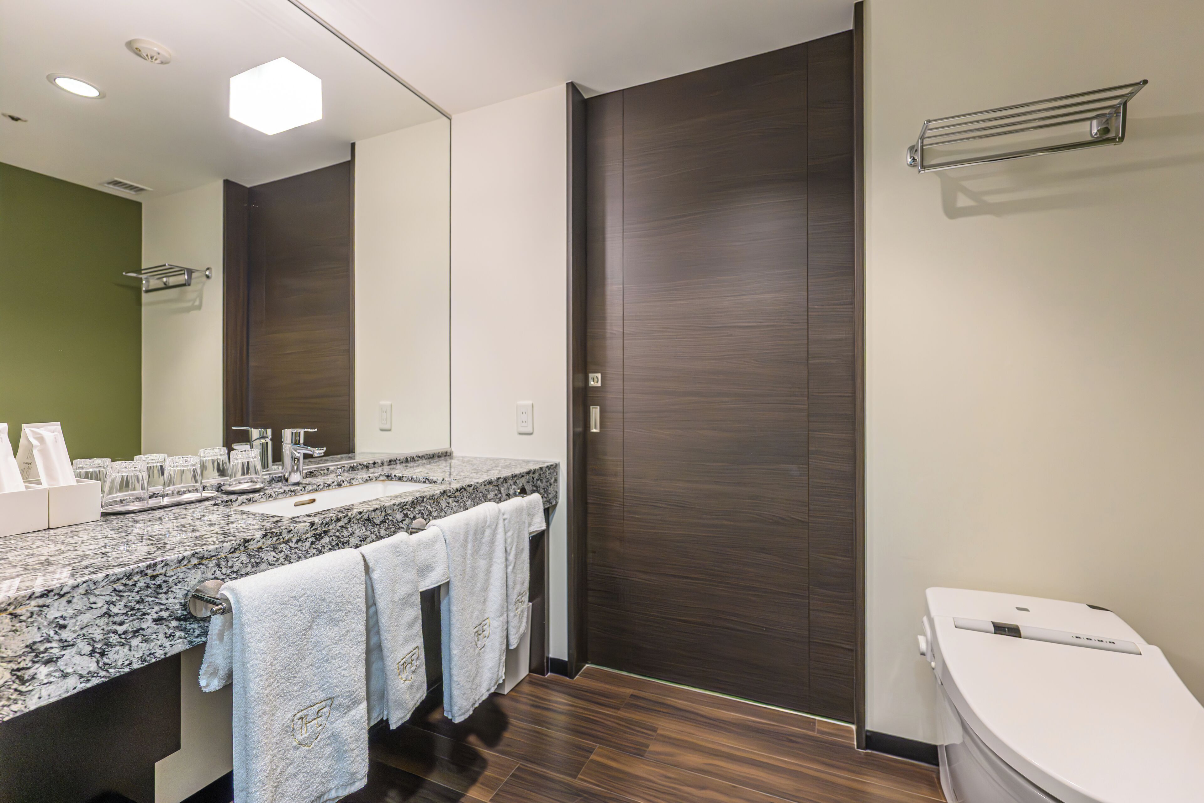A modern bathroom with a granite countertop, a large mirror, a dark wooden door, and a white toilet.
