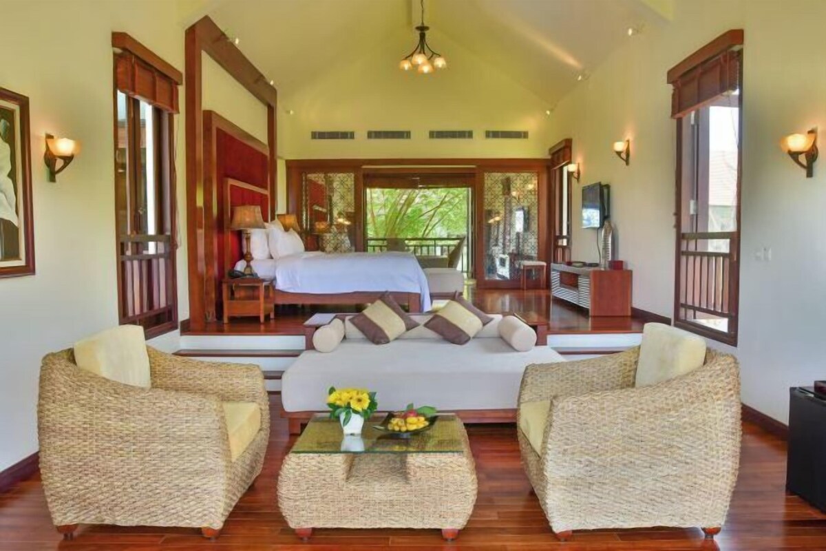 A spacious room with a bed, two wicker sofas, a glass coffee table with flowers, and a view of the outdoors.