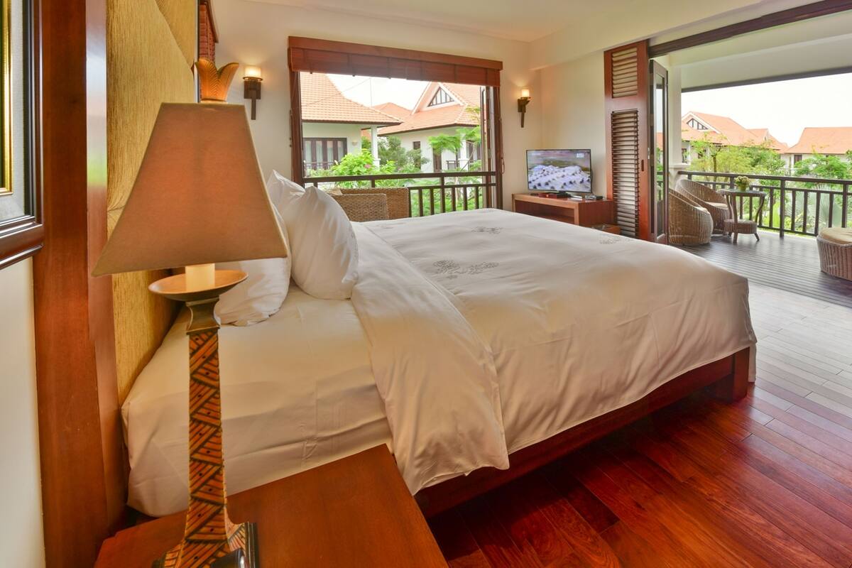 A bedroom with a large bed, a TV, a balcony with wicker chairs, and a view of buildings.