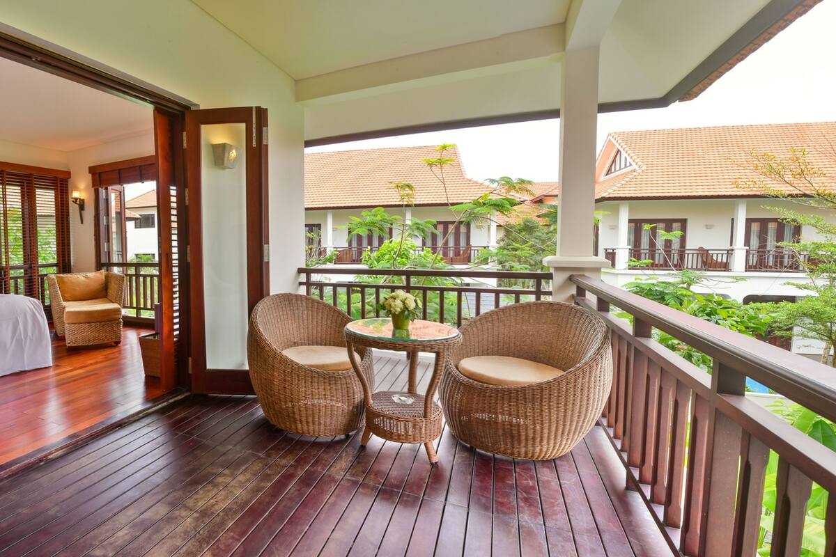 A hotel room with a wooden floor, a bed, a sitting area with wicker chairs, and a balcony with a view of other buildings and greenery.