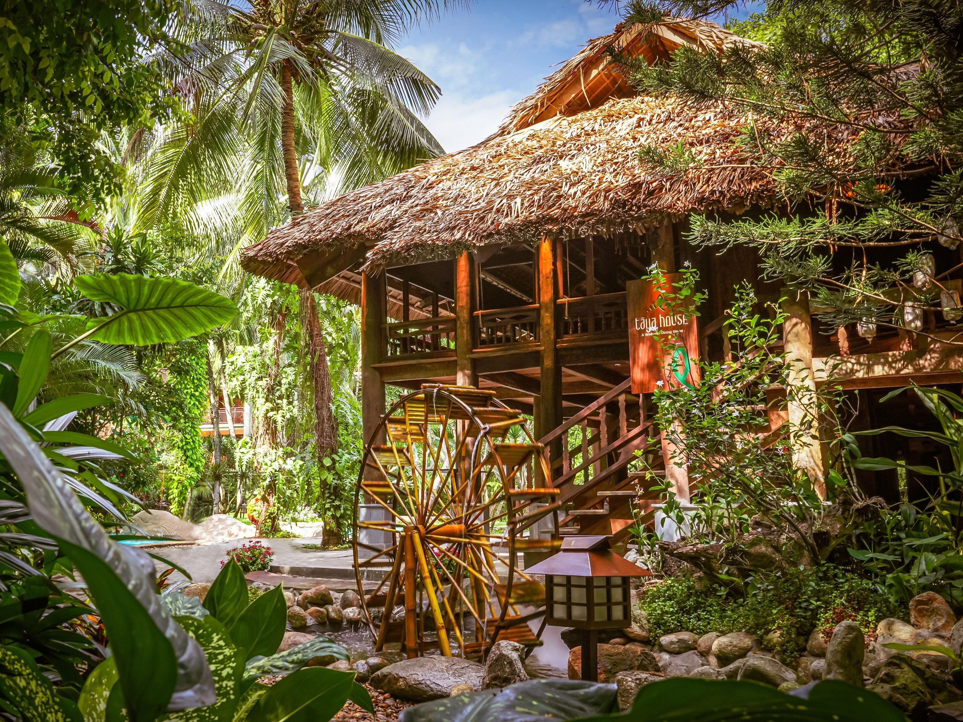 Une construction traditionnelle à toit de chaume, dotée d’un escalier en bois menant à une plateforme, le tout entouré d’une végétation tropicale luxuriante.
