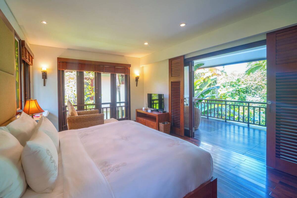 A bedroom with a large bed, a TV, and a balcony with a view of greenery.