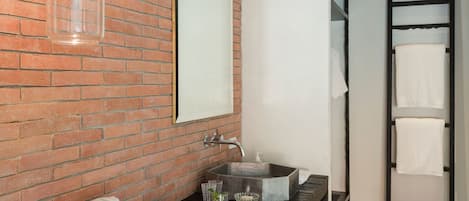 Un baño con pared de ladrillo, un espejo grande, un lavamanos con dispensador de jabón y un perchero para toallas.