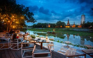 Een buiten eethoek met houten tafels en stoelen, een rivier en een tempel op de achtergrond.