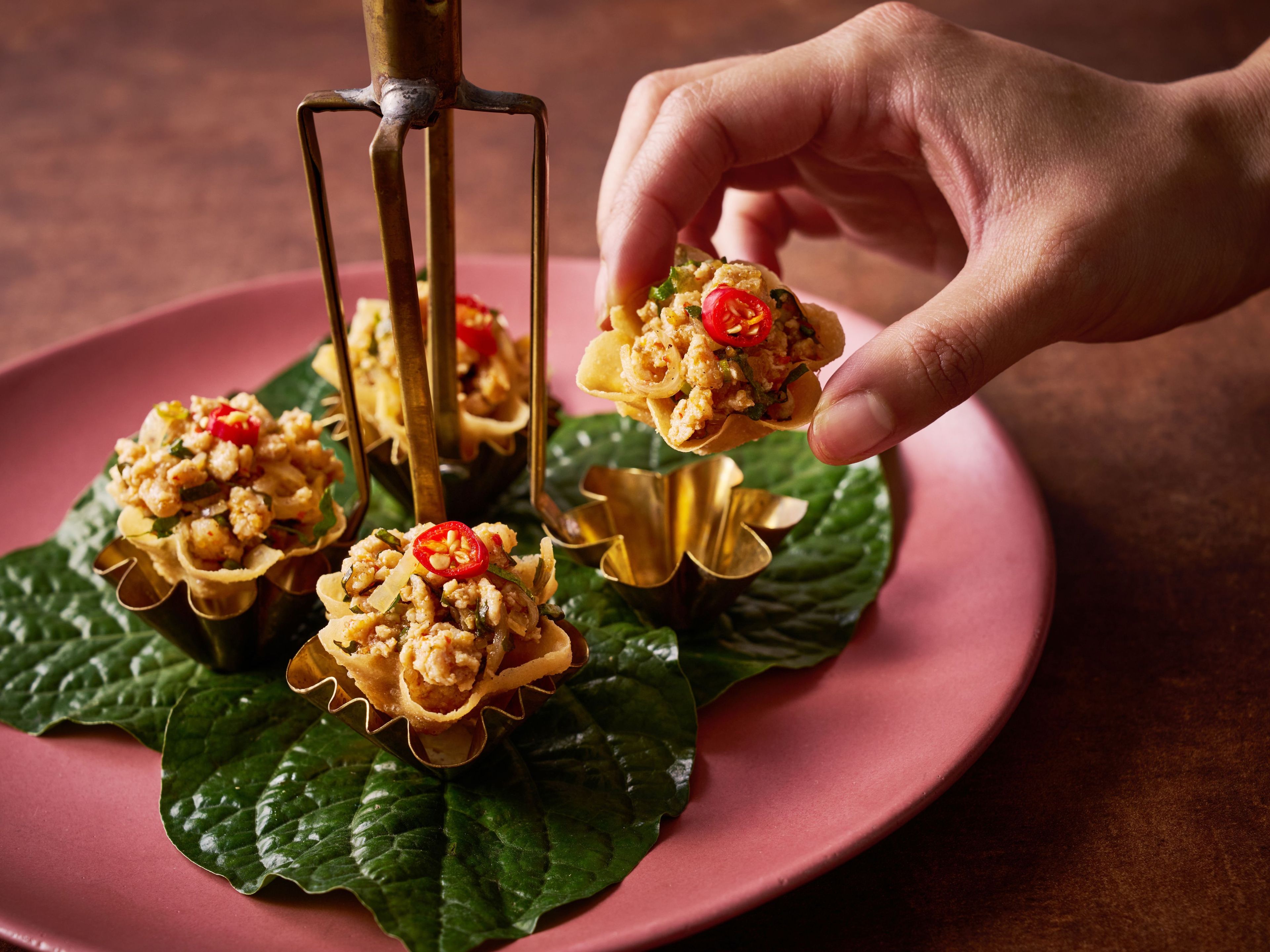 A hand holding a food item with a golden utensil, placed on a pink plate with green leaves underneath.
