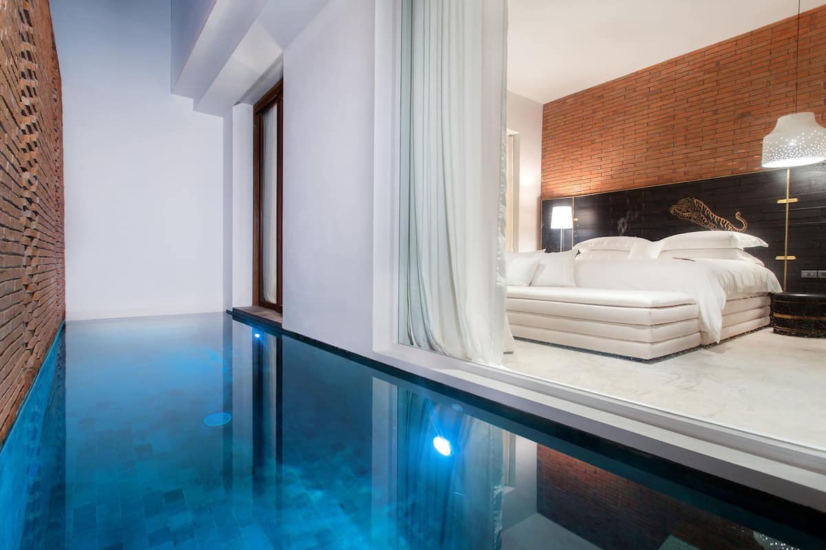 A modern bedroom with a glass-enclosed pool, a brick wall, and a large bed with white bedding.