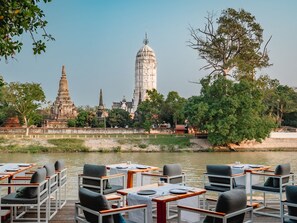 A riverside dining area with tables and chairs, a view of ancient temple structures, and a clear sky. - Sala Ayutthaya (Ayutthaya)