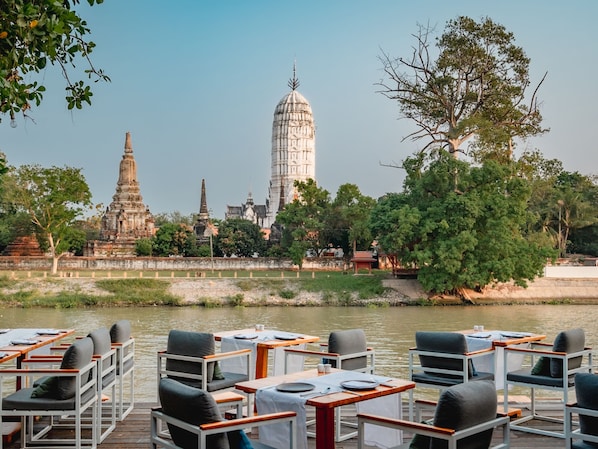 A riverside dining area with tables and chairs, a view of ancient temple structures, and a clear sky. - Sala Ayutthaya (Ayutthaya)