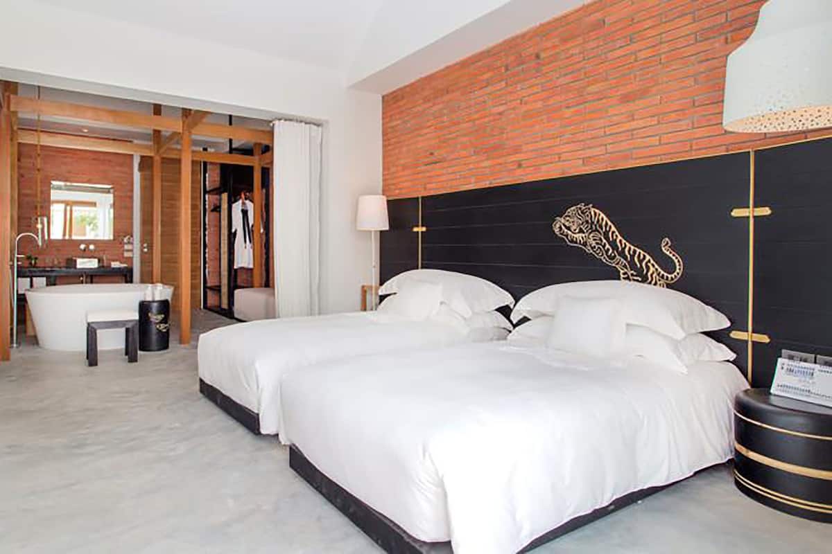 A modern hotel room with a large bed, a flat-screen TV, a bathroom with a bathtub, and a brick accent wall.
