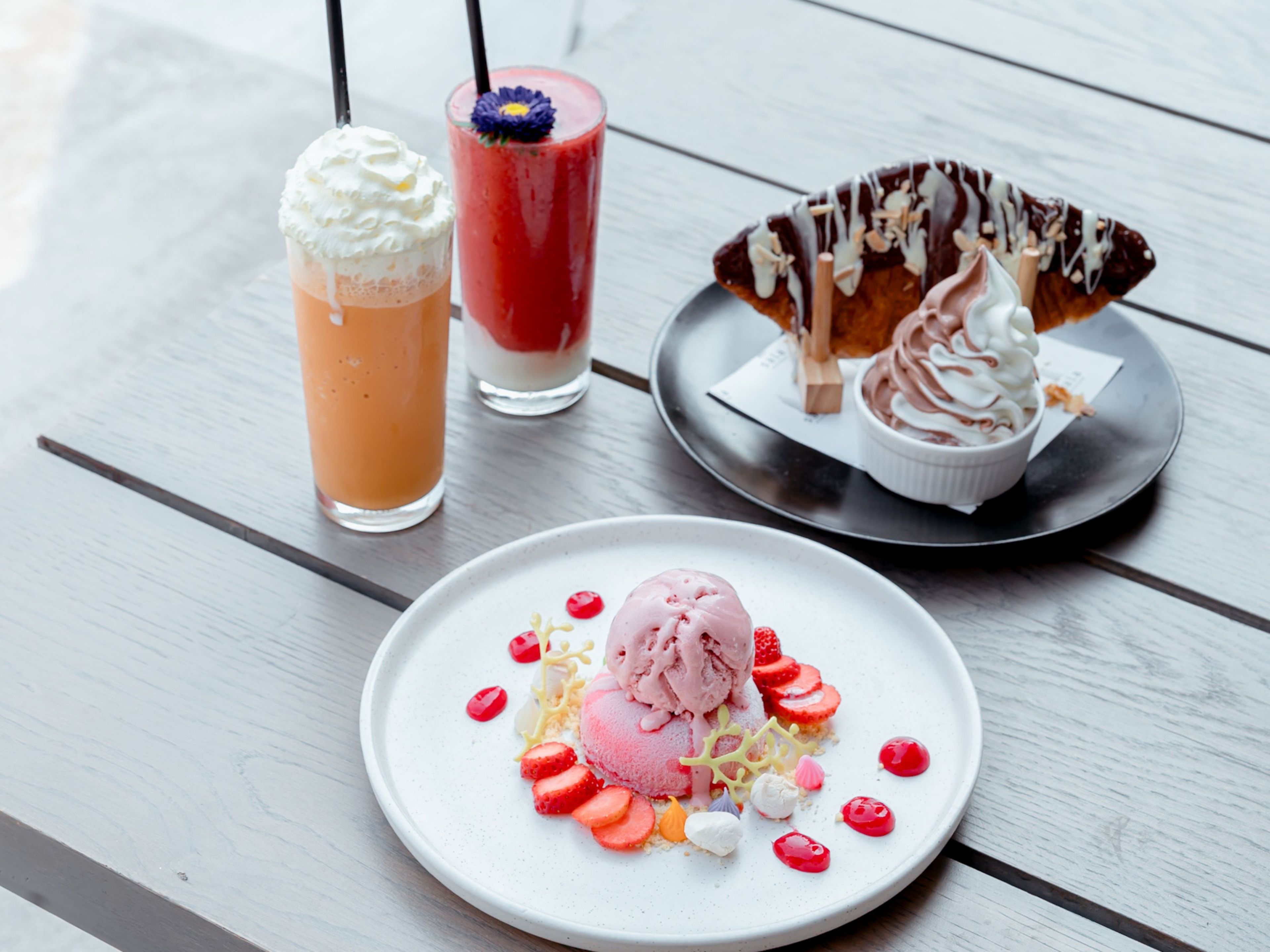 Une assiette de dessert avec de la crème glacée et des fraises, deux boissons garnies de crème fouettée et une pâtisserie nappée de chocolat.