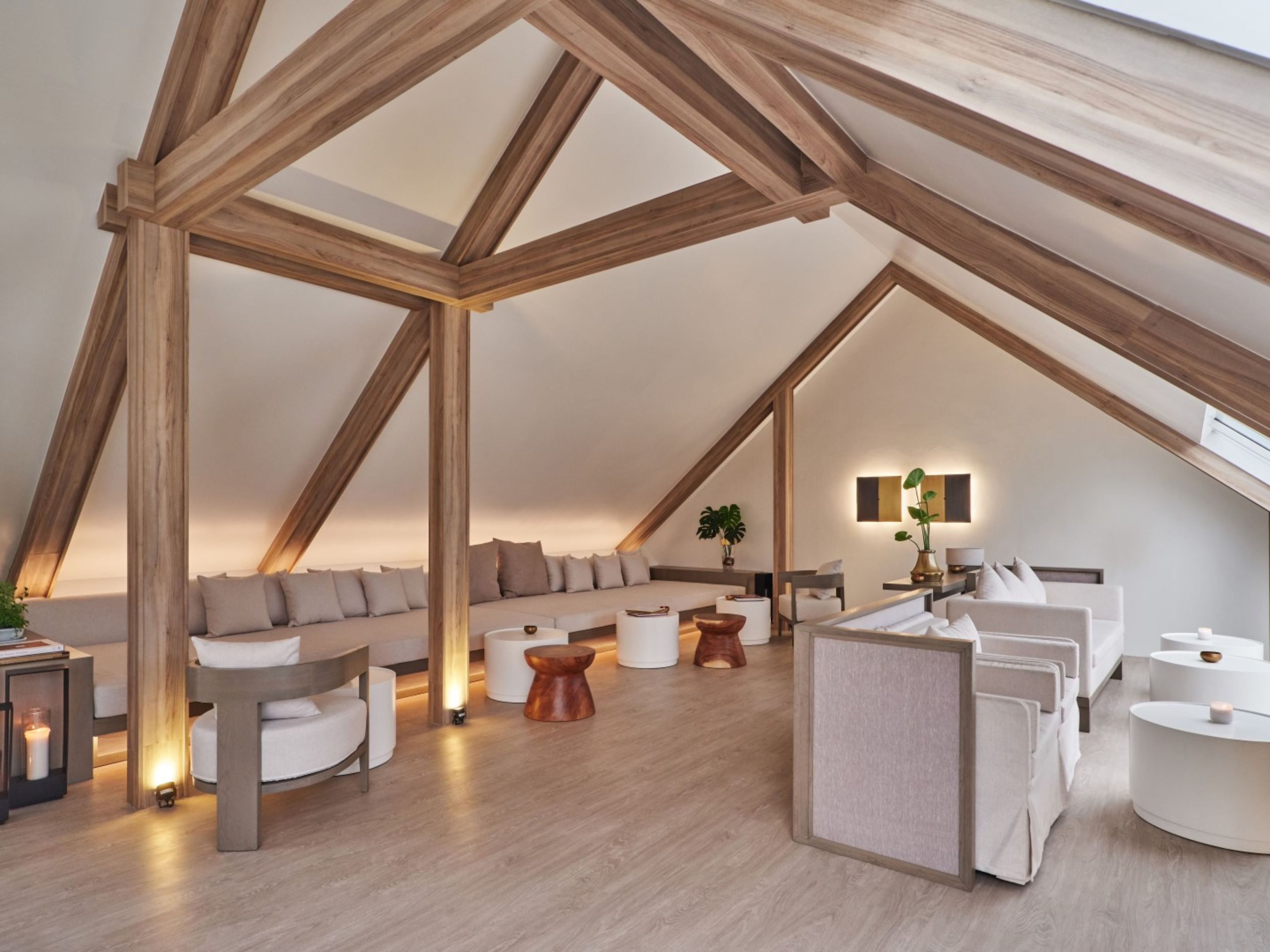 A modern loft bedroom with exposed wooden beams, a large bed, a seating area with sofas, and a small table with a plant.