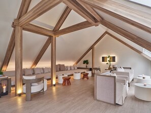 Un dormitorio estilo loft moderno con vigas de madera a la vista, una cama grande, una zona de estar con sofás y una mesita con una planta.
