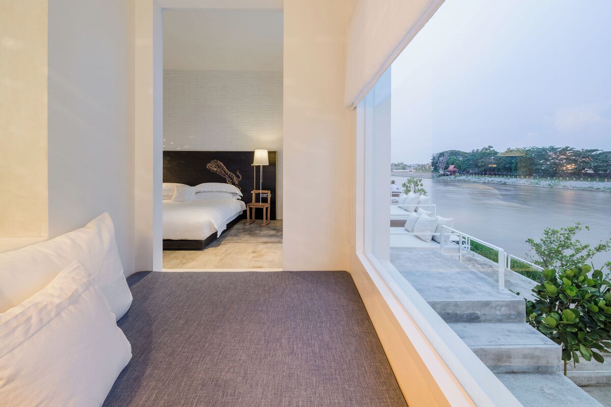 A modern hotel room with a bed, a nightstand, and a lamp. There is a view of a body of water and a cityscape through large windows.