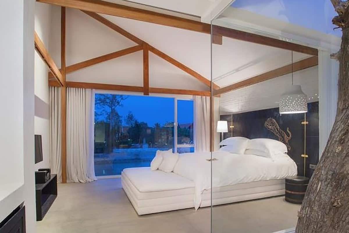 A modern bedroom with a large bed, a glass partition, and a view of the outdoors.