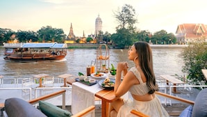 Un comedor con mesas y sillas con vistas a un río, con un bote y estructuras de templo al fondo.