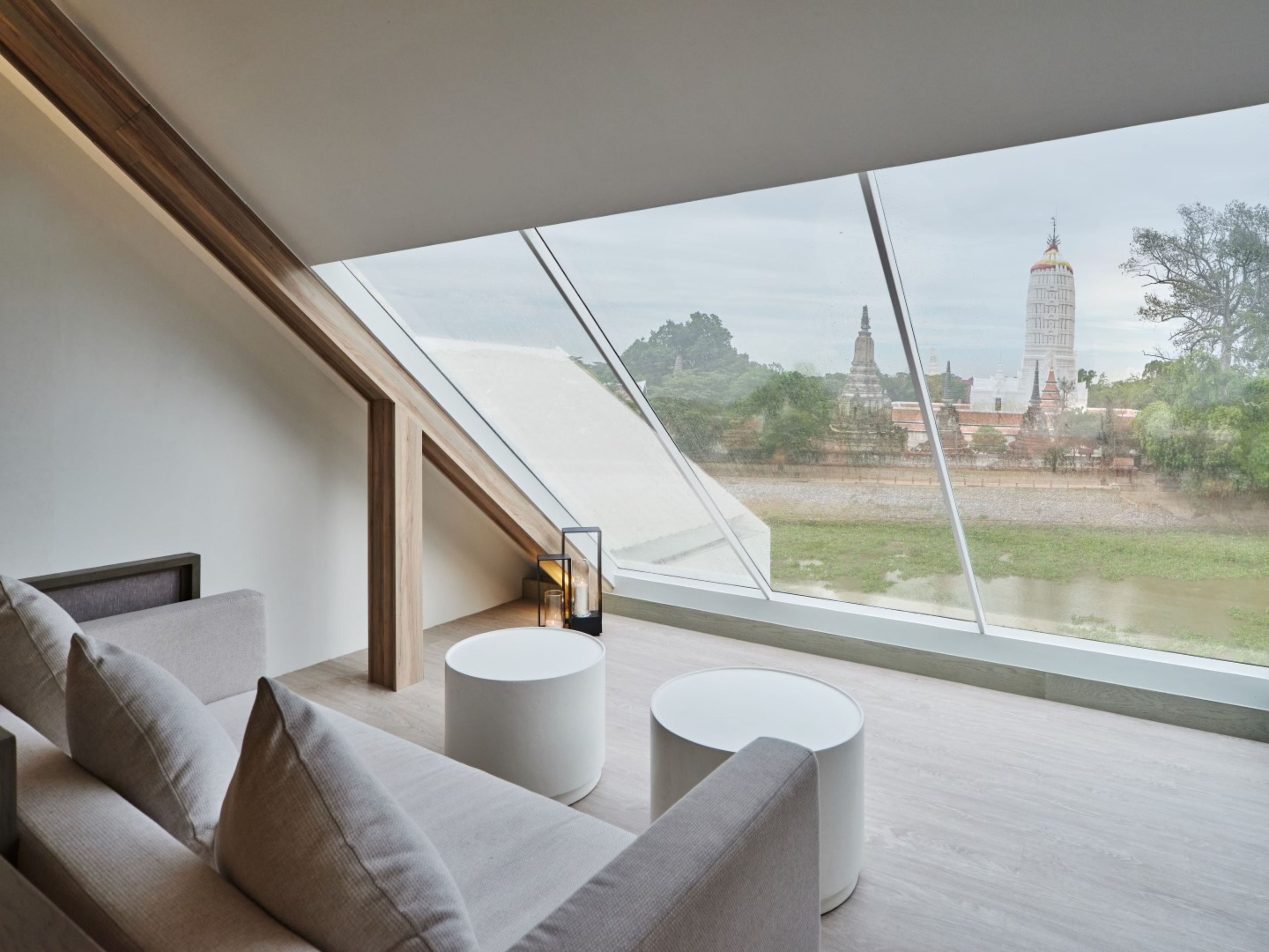 A modern interior with a sloped ceiling, a sofa, two round tables, and a view of a temple through large windows.