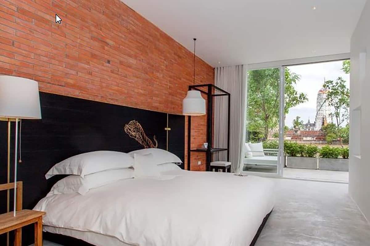 A modern hotel room with a large bed, a brick accent wall, a large window with a view of a temple, and a hanging light fixture.