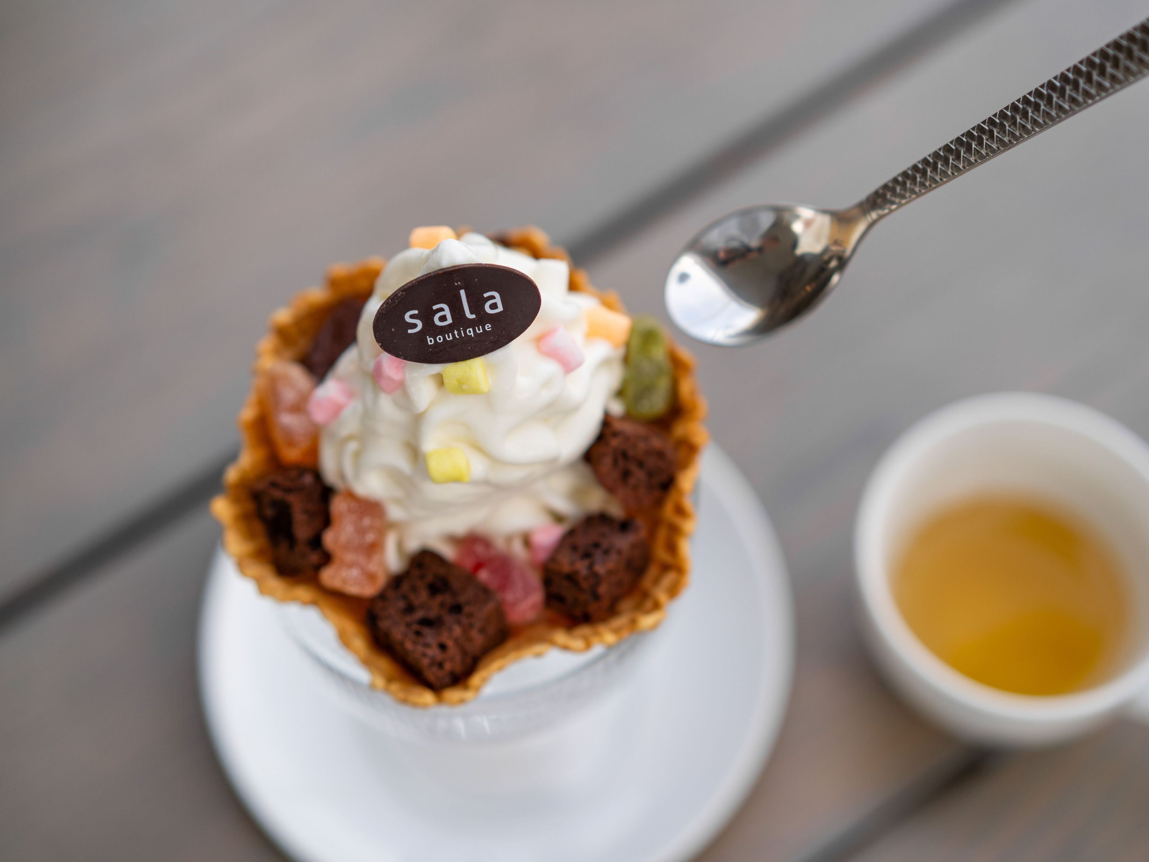 Un postre con una etiqueta que dice 'sala boutique' sobre una cuchara, una taza de líquido amarillo a un lado, y un mango de cuchara con textura.